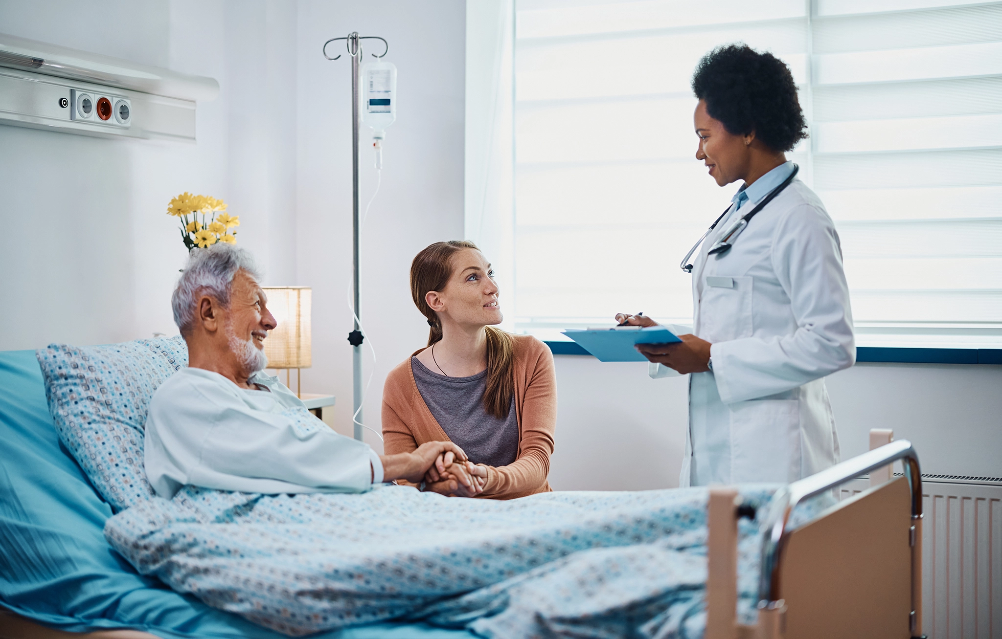 Doctor talking to a patient in a hospital bed and a family member sitting on the side.