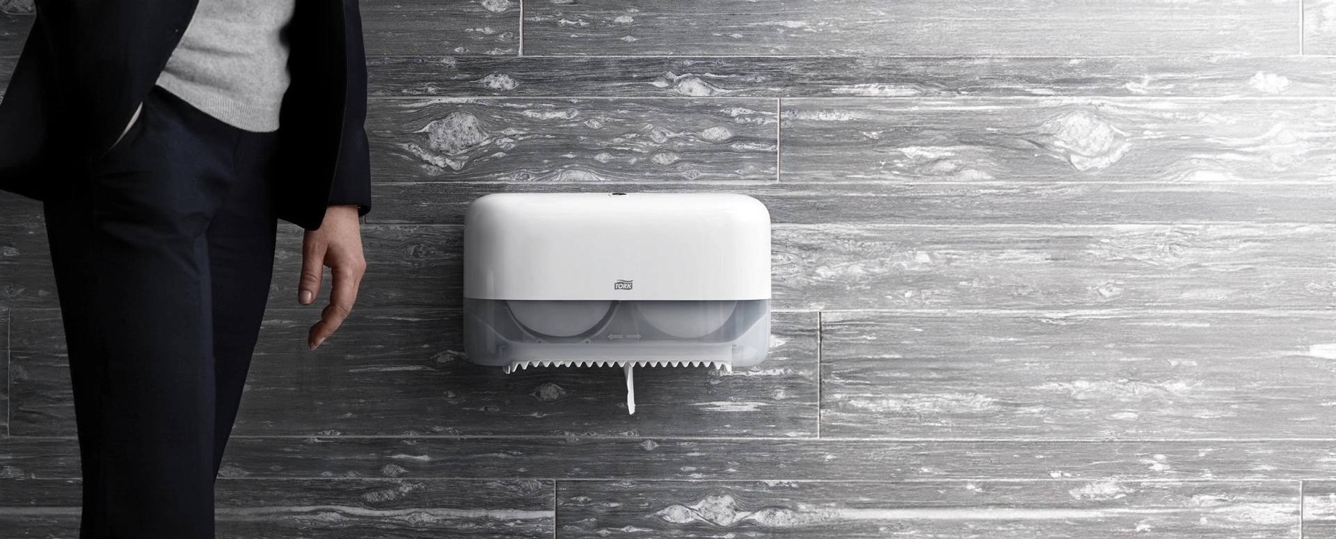 Tork Toilet Paper dispenser mounted on a wall with hands reaching for toilet paper.